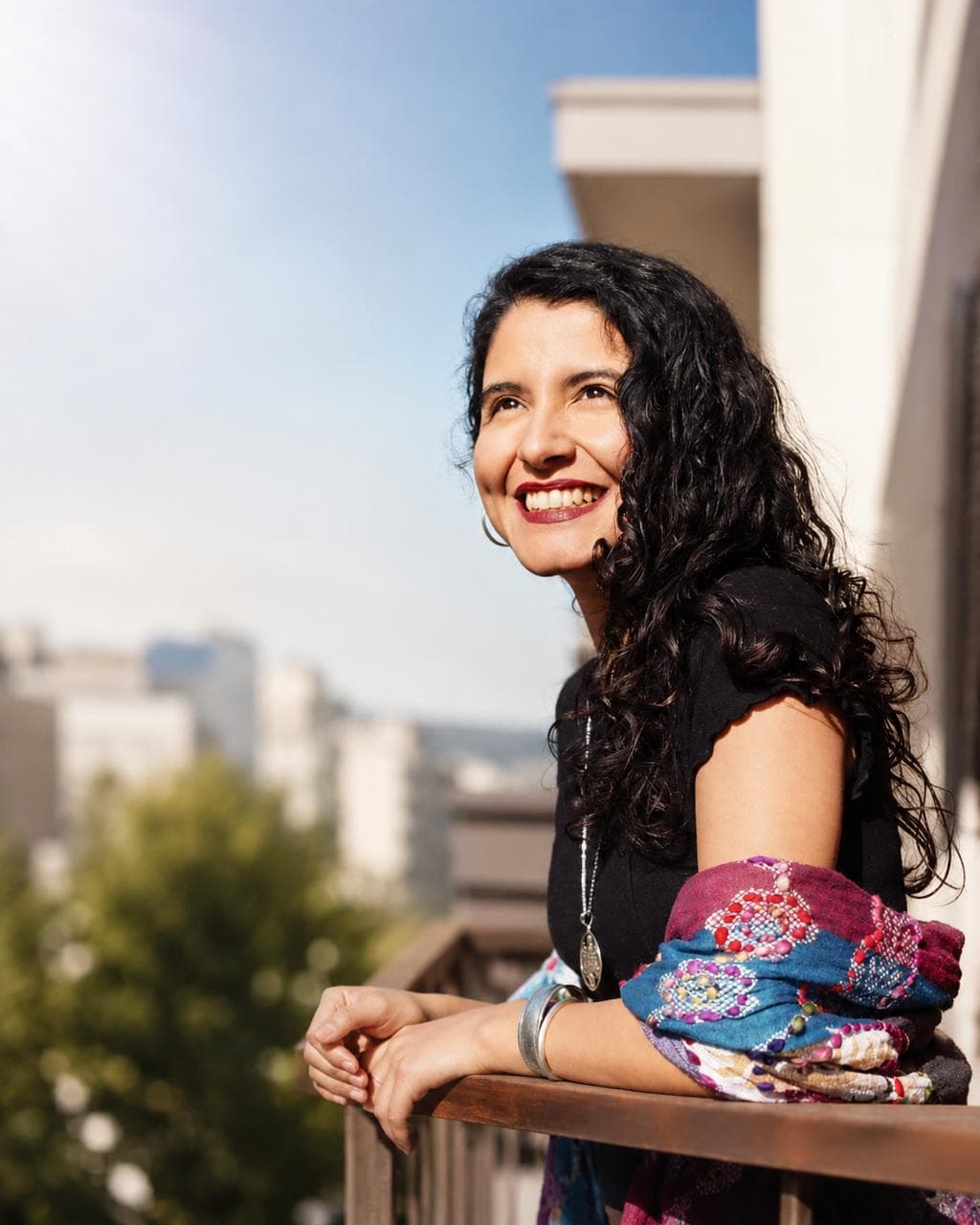 María Alejandra Paredes, sonriendo apoyada en la barandilla de un balcón, con la ciudad al fondo